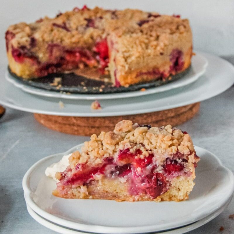 A slice of plum cake sits on two white plates with the remaining cake on white plates on a gray surface.