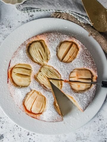 German Sunken Apple Cake sits on a white ceramic plate coated in powdered sugar; being sliced on a light gray surface.