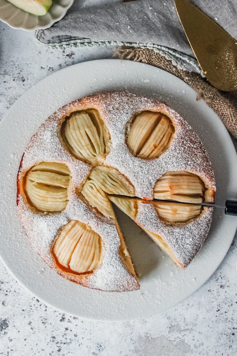 German Sunken Apple Cake sits on a white ceramic plate coated in powdered sugar; being sliced on a light gray surface.