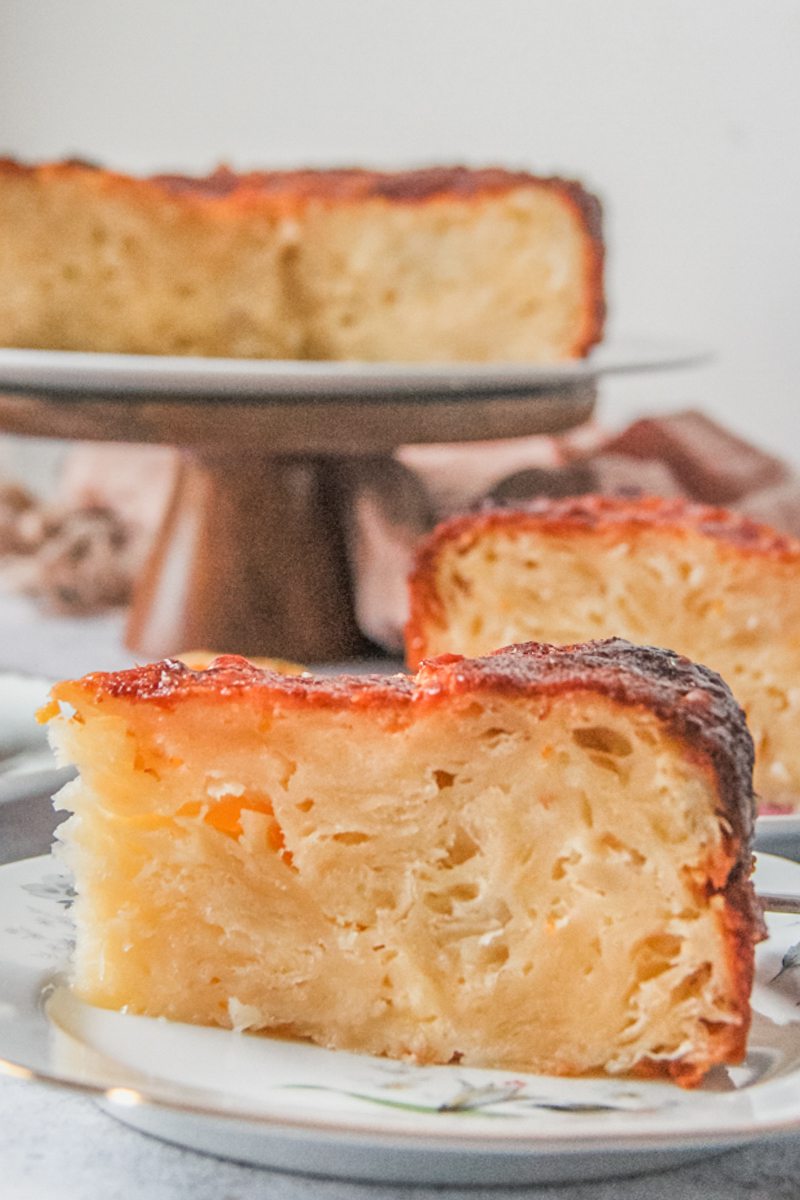 A slice of Greek orange cake sits on an individual plate with another slice behind and the remaining cake on a cake stand on a gray surface.