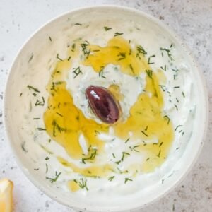 Homemade Greek tzatziki sits in a white ceramic bowl on a light gray surface garnished with a traditional black olive.