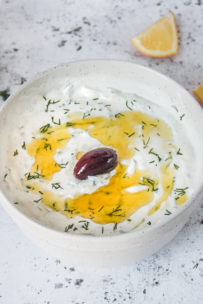 Homemade Greek tzatziki sits in a white ceramic bowl on a light gray surface garnished with a traditional black olive and wedge of lemon on the side.