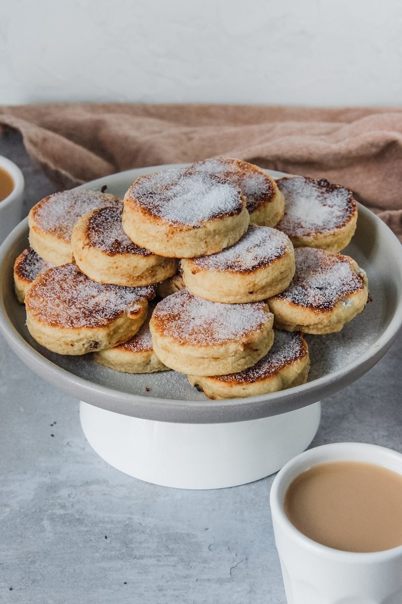 A stack of Welsh cakes sprinkled with sugar sit on a rimmed ceramic plate with a cup of tea visible in the foreground.