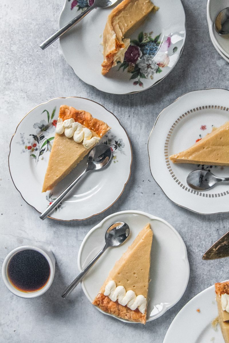 Individual slices of butterscotch pie sit on white ceramic plates on a gray surface with a cup of coffee beside.