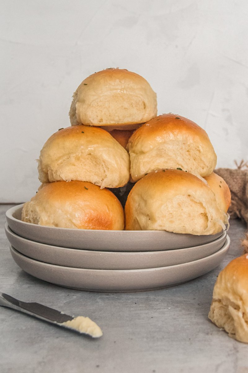 A side view of fluffy soft dinner rolls stacked on a stack of ceramic plates on a gray surface.