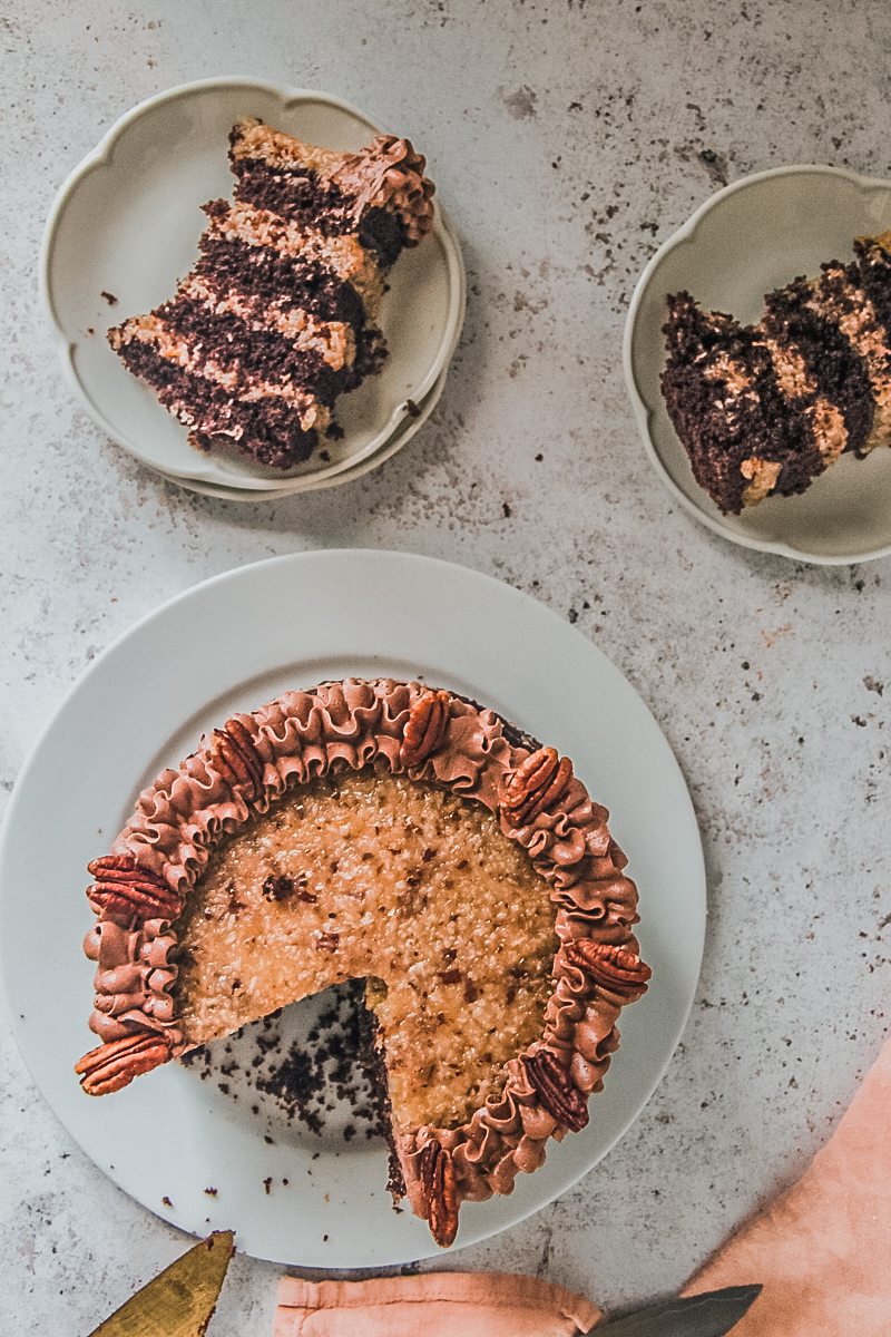 Two slices of German chocolate cake lay on fluted white plates next to the cake on a light gray surface.