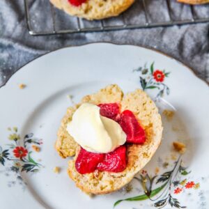 A halved scone topped with roasted strawberries and lightly whipped cream sit on a floral individual plate on a gray cloth.