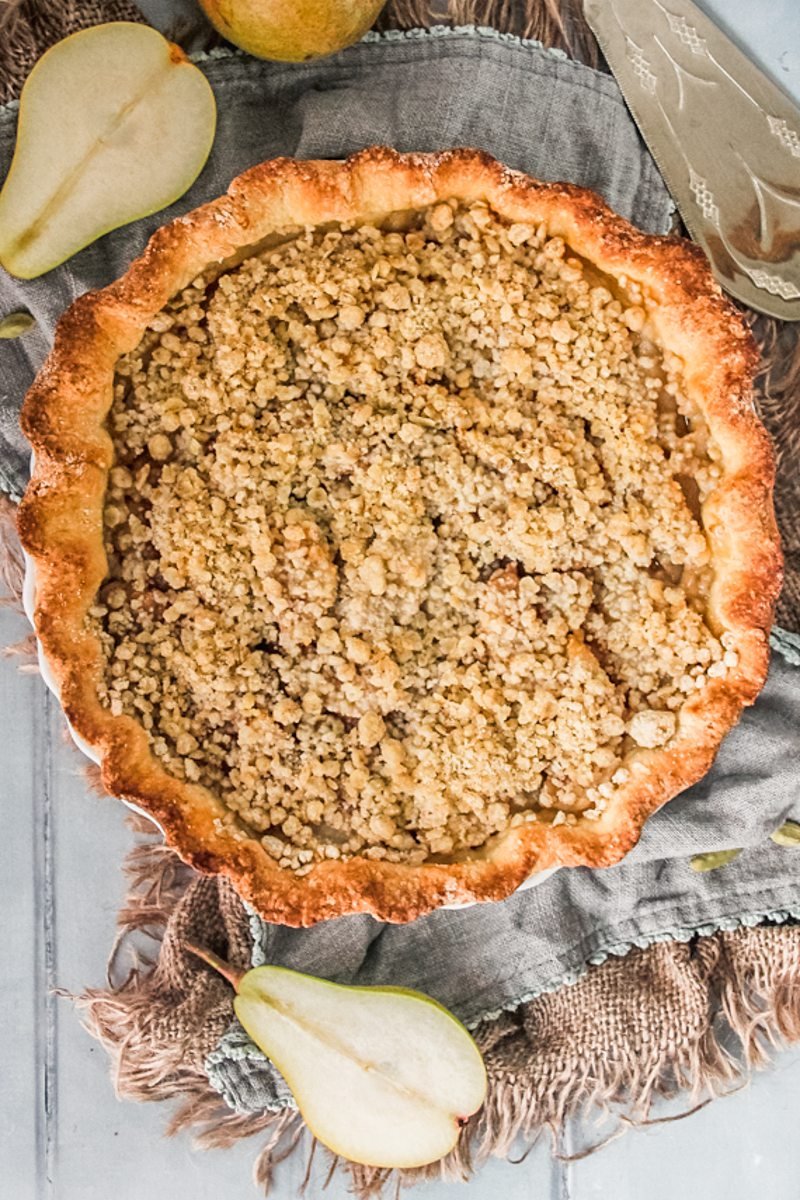 A pear streusel pie sits on top of two cloths on a blue gray surface with halved pears beside.