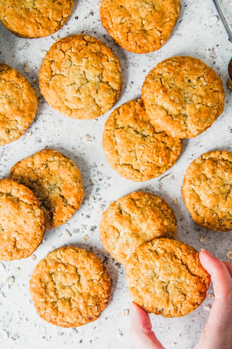 An Anzac biscuit is picked up from a light gray surface with other Anzac biscuits beside.