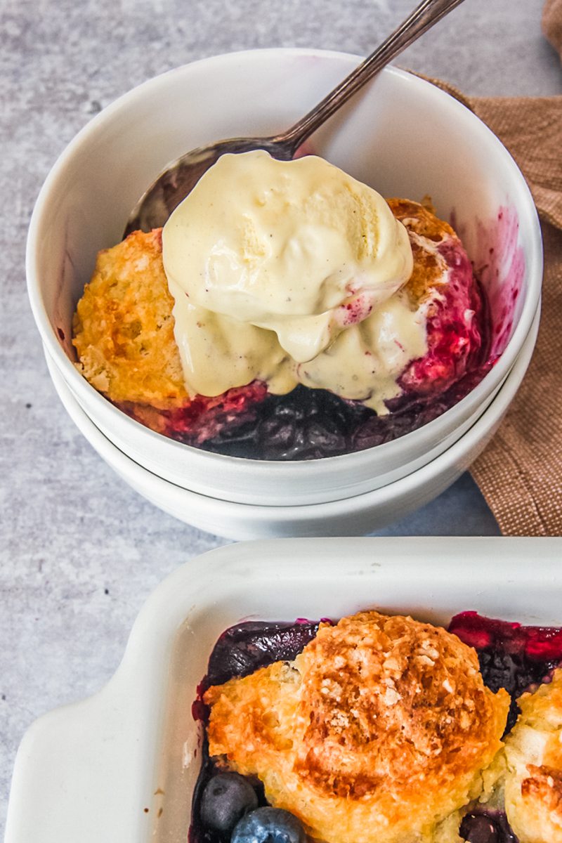 A serving of blueberry cobbler sits in a stack of white ceramic bowls with a scoop of vanilla ice cream on top on a gray surface.