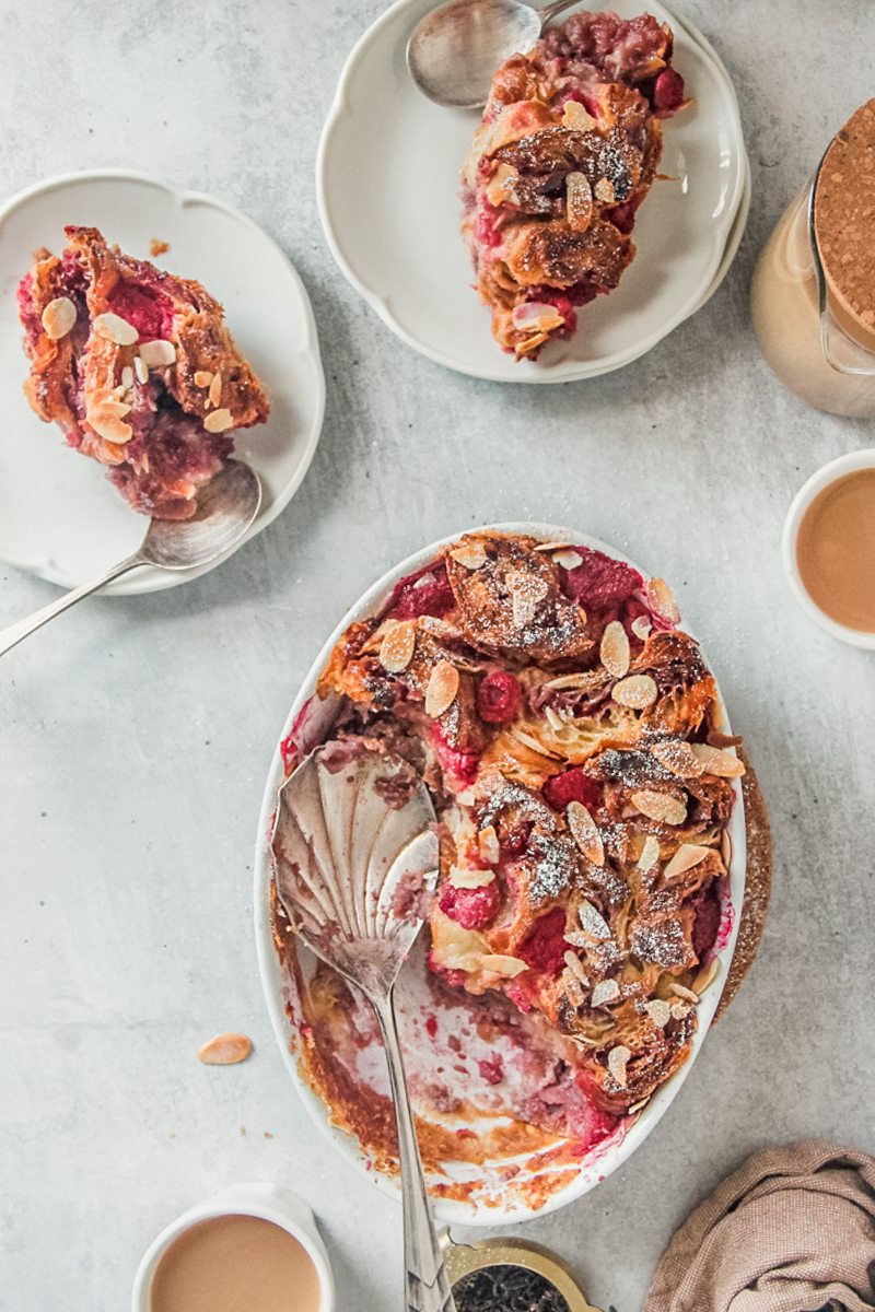 Individual servings of croissant bread pudding sit on white ceramic plates on a gray surface beside an oval white ceramic baking dish.