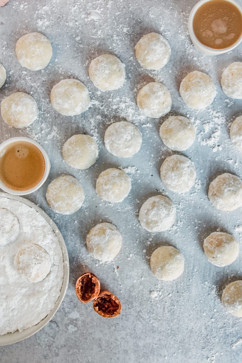 Mexican Butter Cookies also known as Polvorones sit on a gray surface with two cups of coffee and a rimmed ceramic plate with powdered sugar beside.