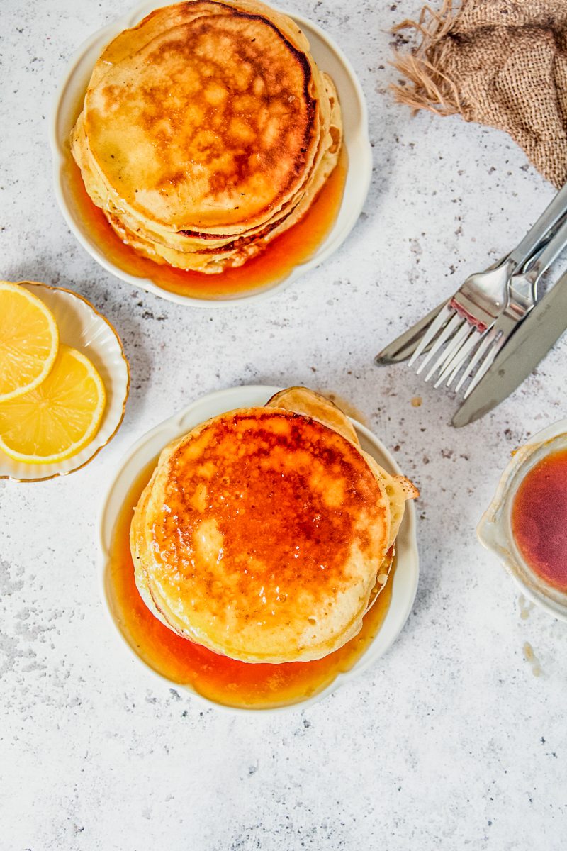 Two stacks of lemon pancakes sits on two individual plates on a light gray surface topped with a lemon sugar syrup.