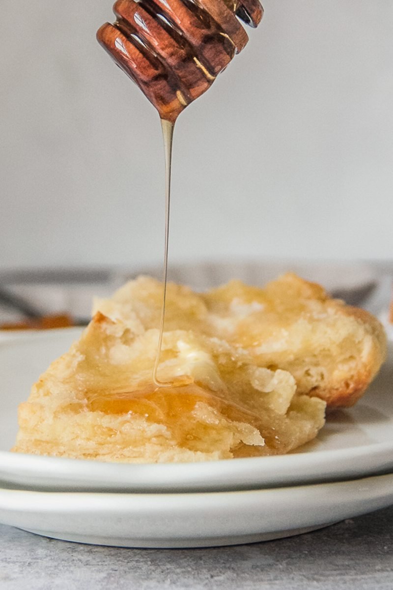 Honey is drizzled over the center of a buttermilk biscuit sitting on a stack of white ceramic plates on a gray surface.