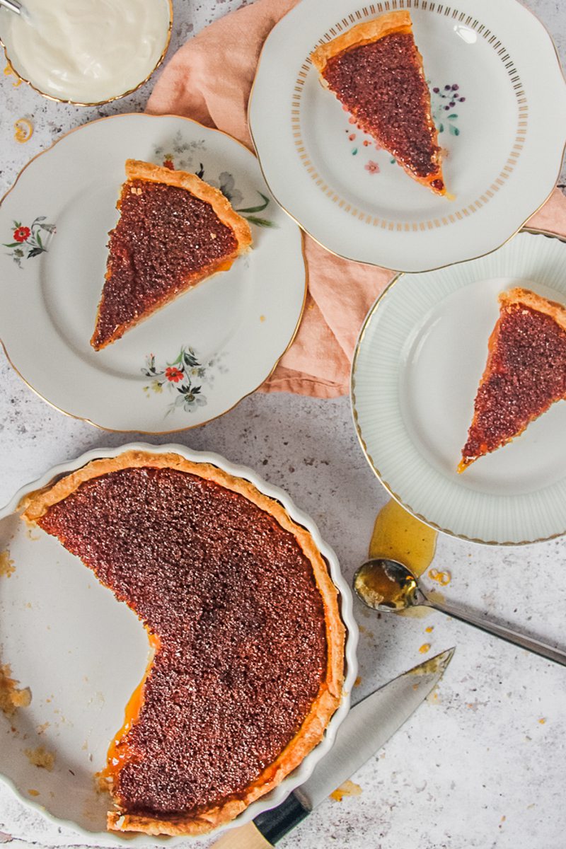 Servings of Treacle Tart sit served up on individual plates beside the remaining tart on a light gray surface.