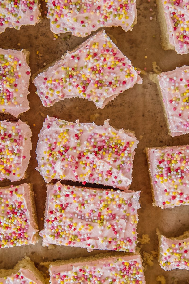 Sugar cookie bars sit on a parchment paper with a raspberry buttercream frosting and hundreds and thousands sprinkles.