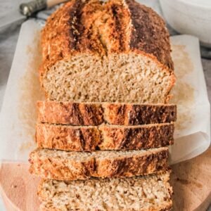 A loaf of Irish Brown bread sits on an oval wooden board with slices laying on the board on a gray surface with a bowl of soup beside.