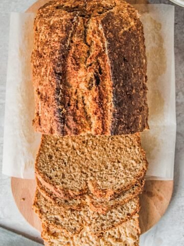 A golden loaf of Irish Brown bread sits on an oval wooden board with slices laying on the board on a gray surface.