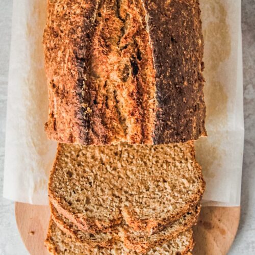 A golden loaf of Irish Brown bread sits on an oval wooden board with slices laying on the board on a gray surface.