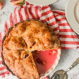 An Irish Rhubarb Pie sits with a few slices removed in a baking dish on a striped cloth on a light gray surface.