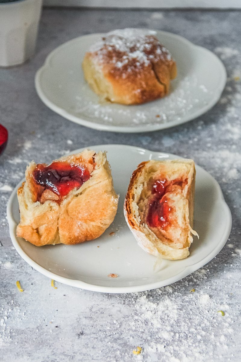 An Austrian jam filled bread roll sits torn in half on a white plate on a gray surface with another roll served up beside.