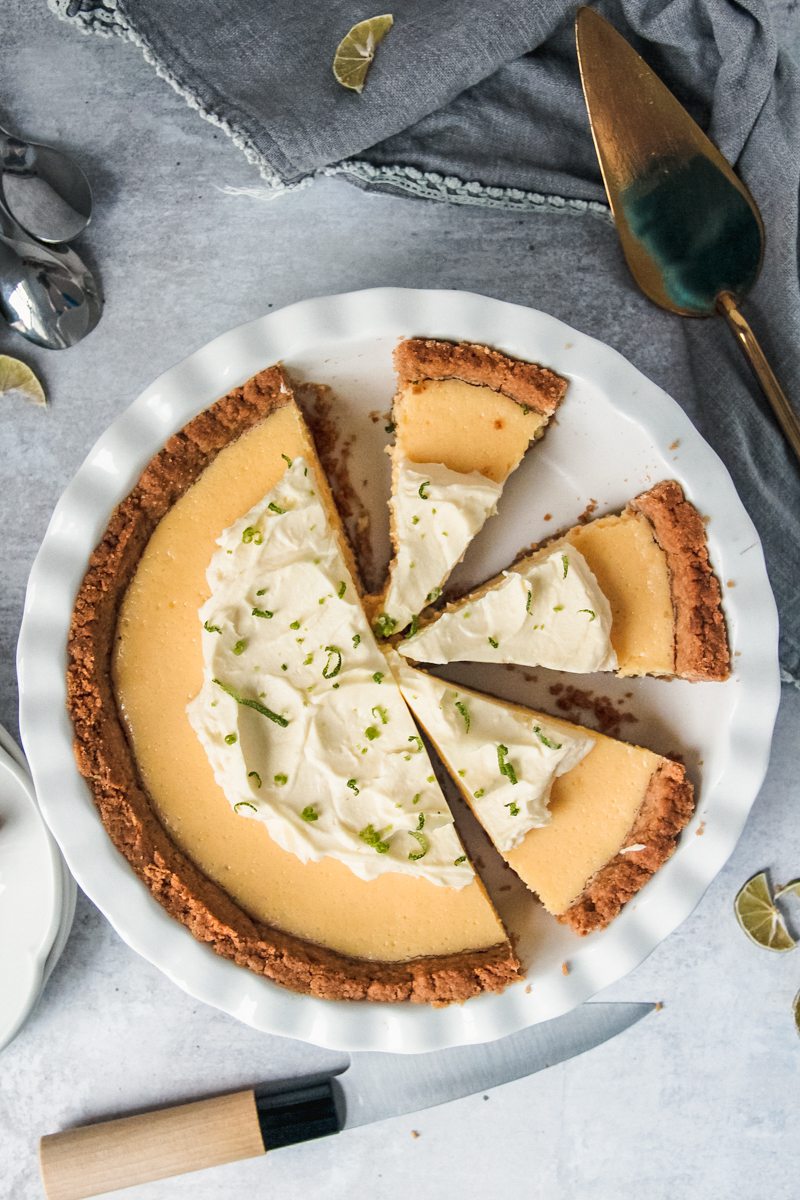 Key Lime pie sits cut in a white ceramic pie dish with three pieces cut and ready to be served on a gray surface.