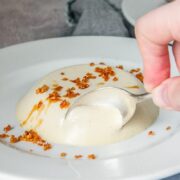 A spoon is dug into an individual serving of manuka and cream panna cotta on a white ceramic plate on a gray surface.