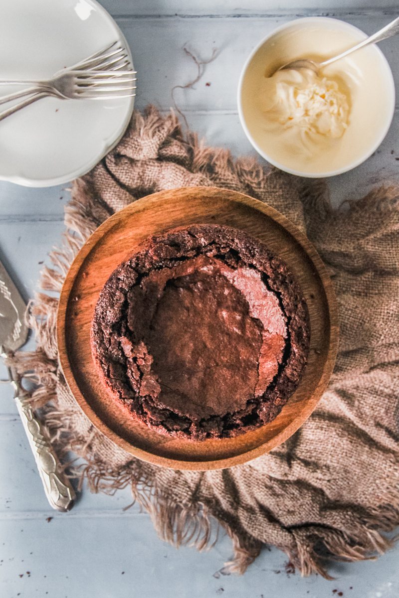 A Kladdkaka or Swedish Chocolate Cake sits on a wooden cake stand with a bowl of whipped cream and a stack of white ceramic plates beside on a light blue surface with a brown cloth beside.