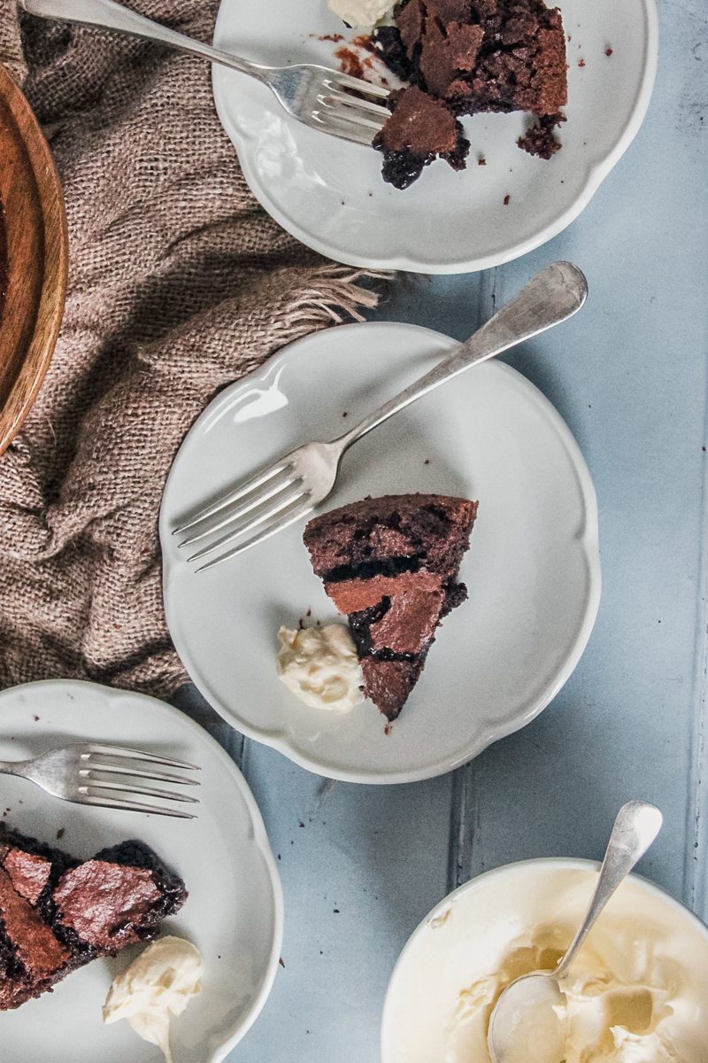 Slice sof Fudgy Swedish Chocolate cake are served up on an individual white ceramic plate with it's gooey interior visible beside a dollop of whipped cream on a light blue surface.