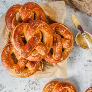 Golden pretzels flecked with pieces of sea salt sit stacked on a plate and a couple on a light gray surface.