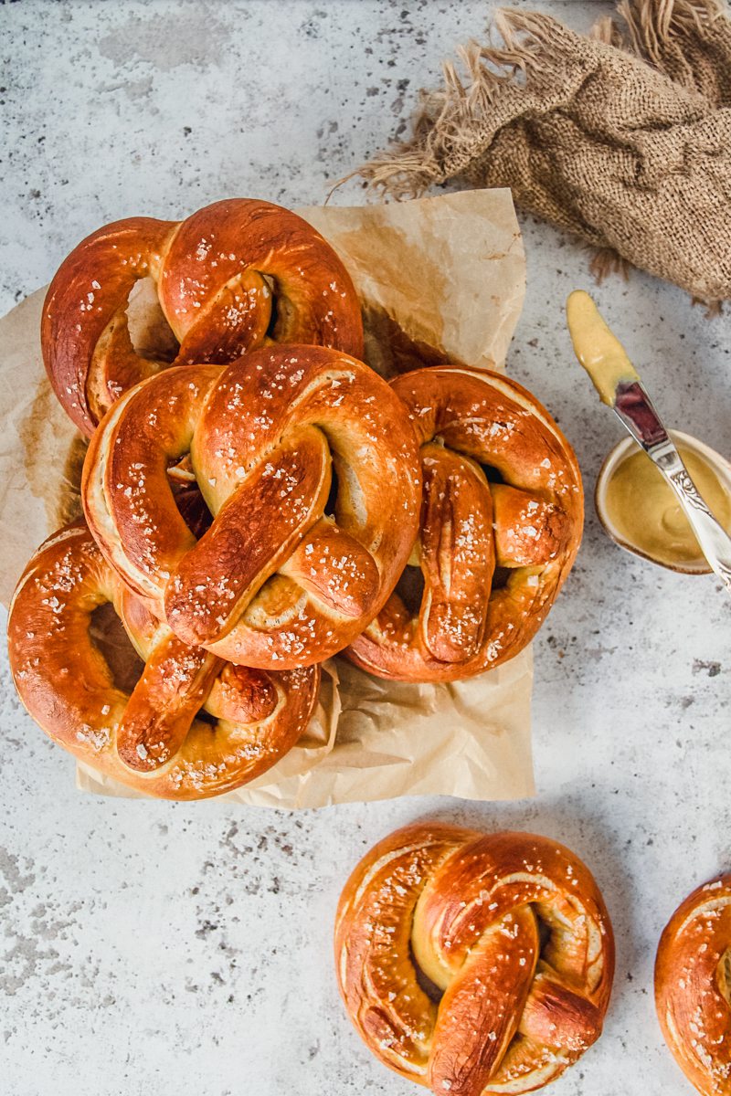 Golden pretzels flecked with pieces of sea salt sit stacked on a plate and a couple on a light gray surface.