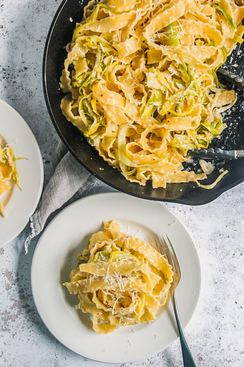 A cast iron skillet with leek pasta topped with grated parmesan sits on a light gray surface with an individual white ceramic plate.