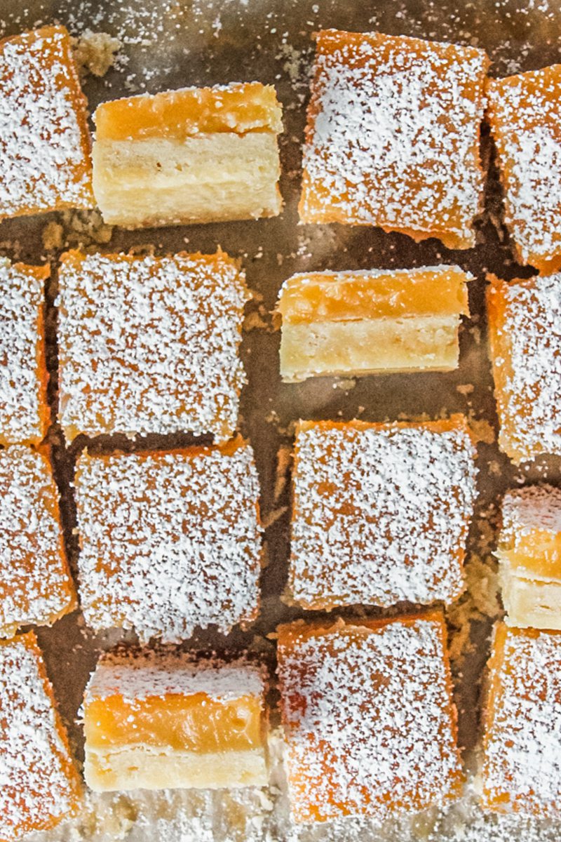 Lemon shortbread bars sit on a parchment paper lined surface with a few sitting on their side showing the shortbread base and lemon curd topping visible.