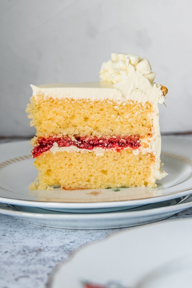 A slice of lemon layer cake with a roasted strawberry center frosted with a chamomile Swiss buttercream frosting sits on a ceramic white plates above a light gray surface.