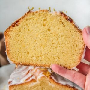 A slice of lemon sponge cake is held up to show the light texture of the cake with the remaining loaf cake behind.