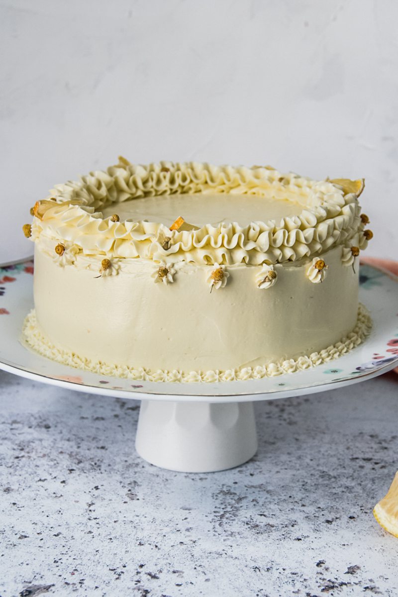 A lemon layer cake with chamomile Swiss meringue frosting sits on a floral ceramic cake stand on a light gray surface.