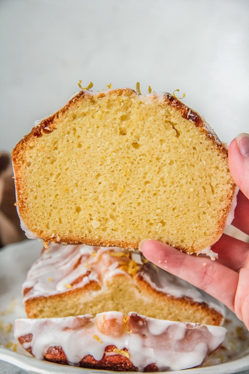 A slice of lemon sponge cake is held up to show the light texture of the cake with the remaining loaf cake behind.