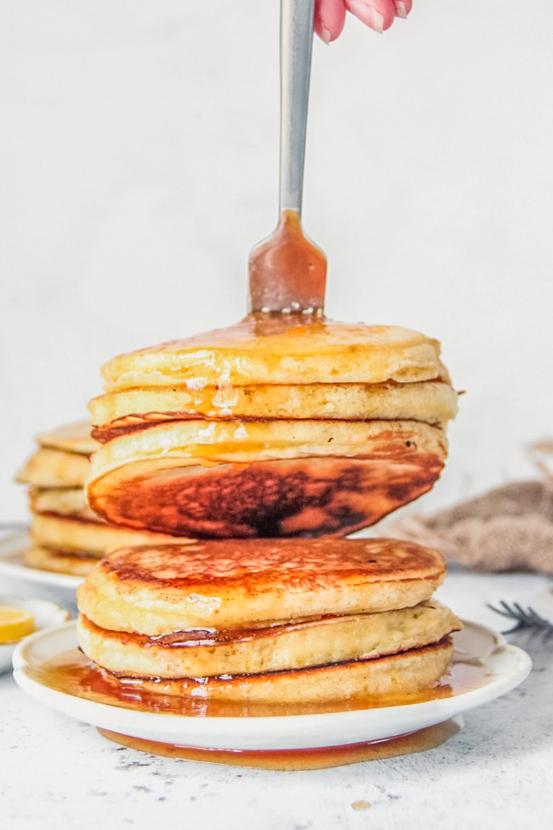 Three lemon pancakes are picked up with a fork from a stack sitting on a white ceramic plate on a light gray surface.