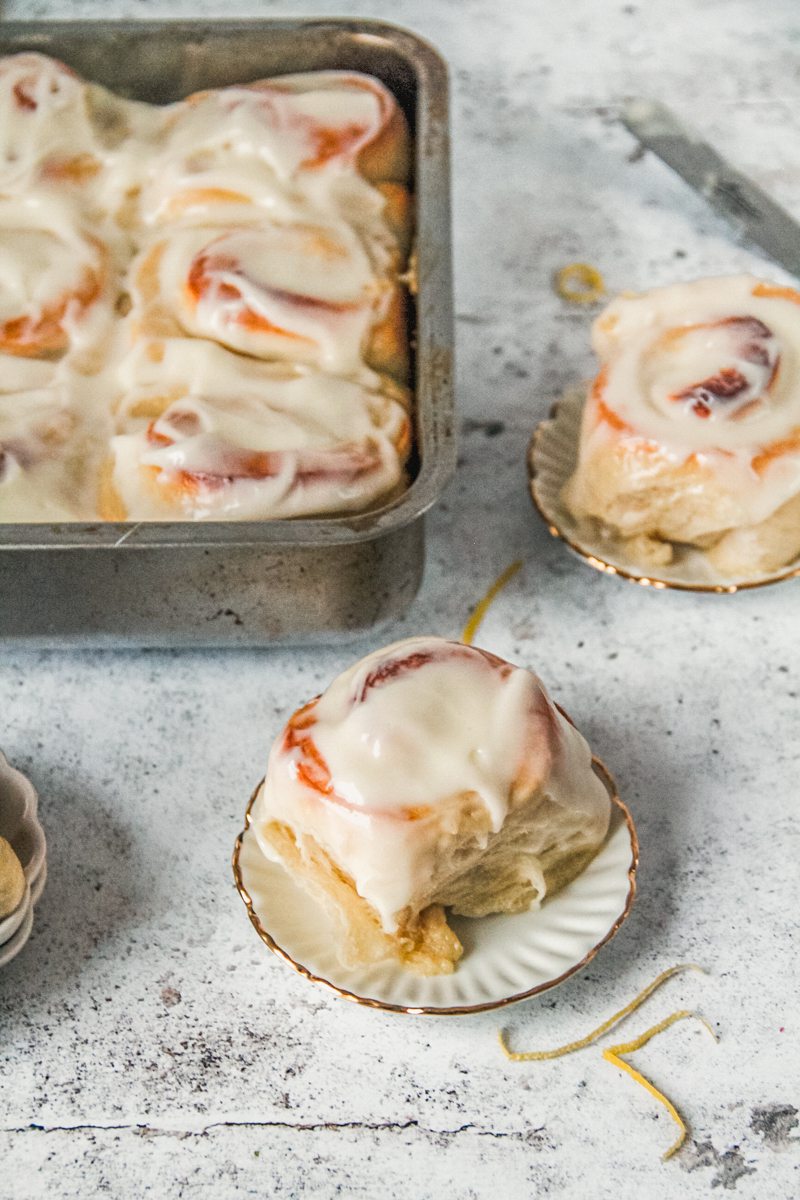 A fluffy lemon roll topped with a lemon cream cheese frosting sits on an individual ceramic plate beside the tin of other lemon rolls.