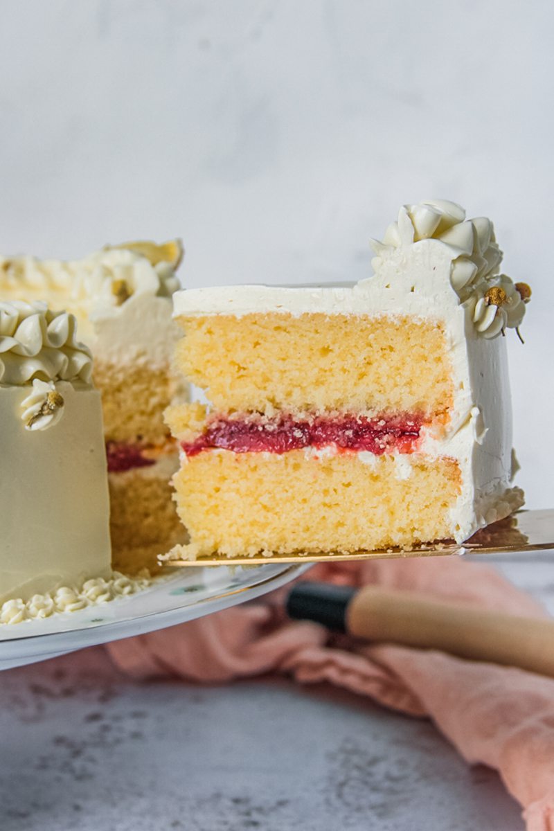 A slice of lemon layer cake with a roasted strawberry center frosted with a chamomile Swiss buttercream frosting sits on a cake server above a light gray surface.