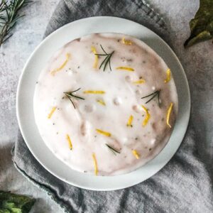 A lemon cake with rosemary glaze sits on a white ceramic plate on a gray cloth on a gray surface.