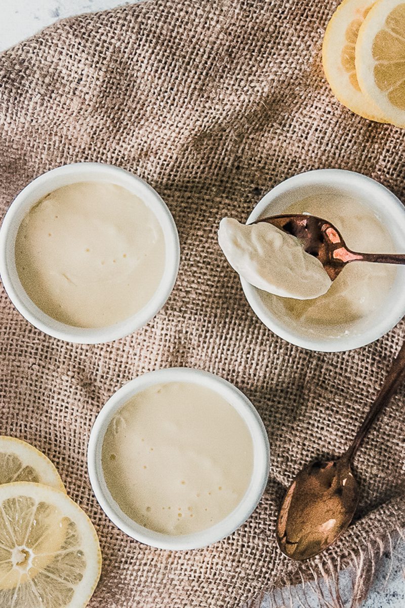 Individual servings of lemon posset sit in white ceramic espresso cups on brown cloth with one copper spoonful resting on one serving.