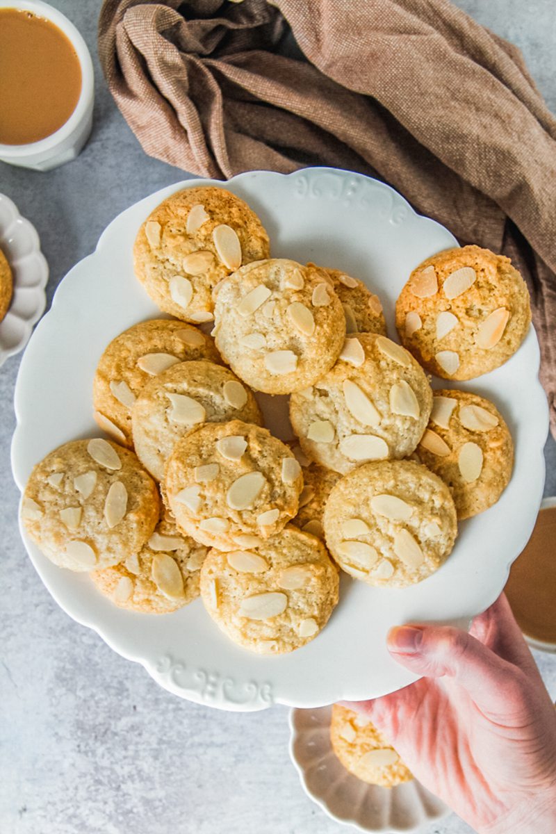 Small golden almond macaroon cookies or Dutch Makroner sit on a fluted white ceramic plate on a gray surface.