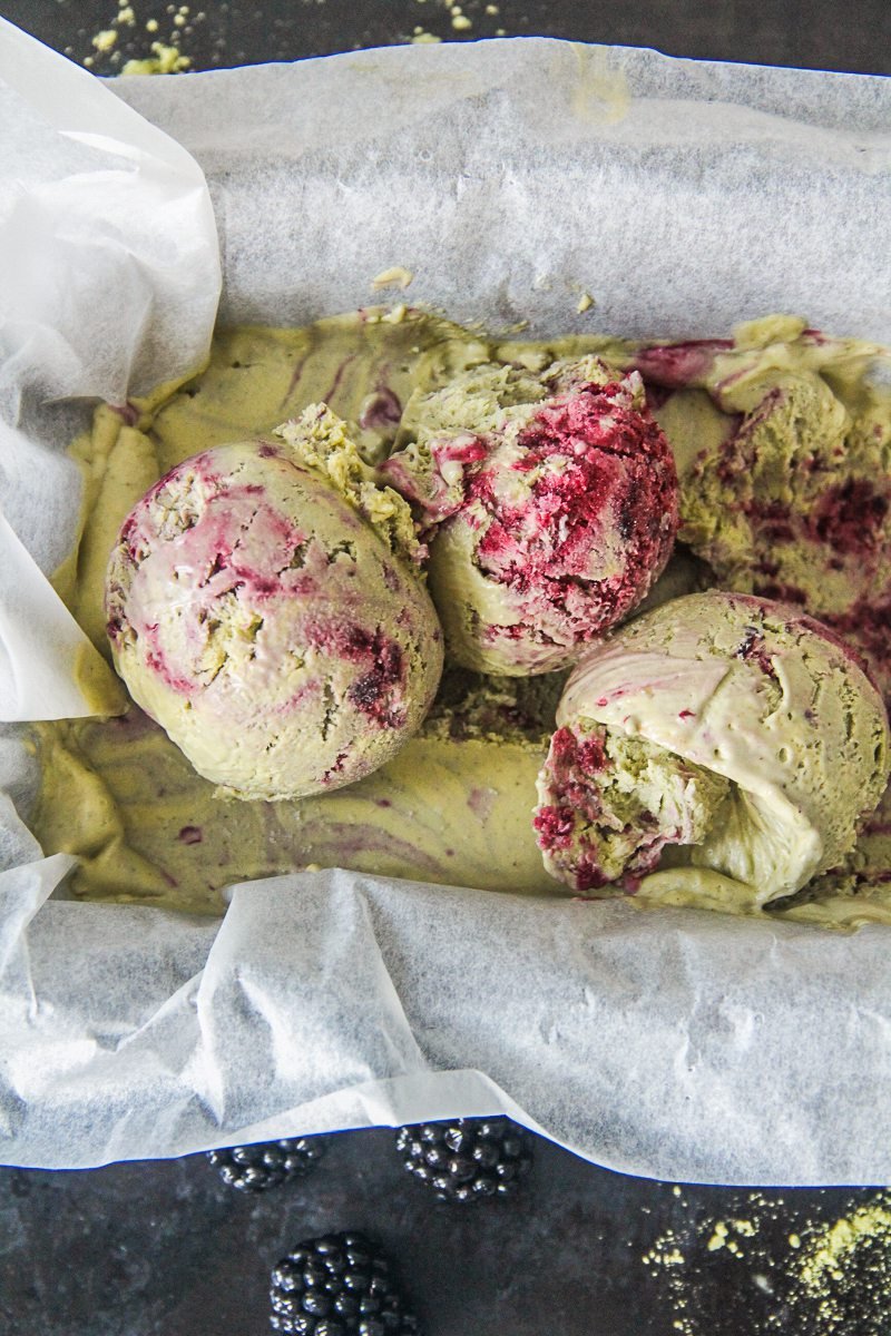 Scoops of matcha blackberry semifreddo sit on top of a tin of semifreddo in a parchment paper lind tin on a dark surface.