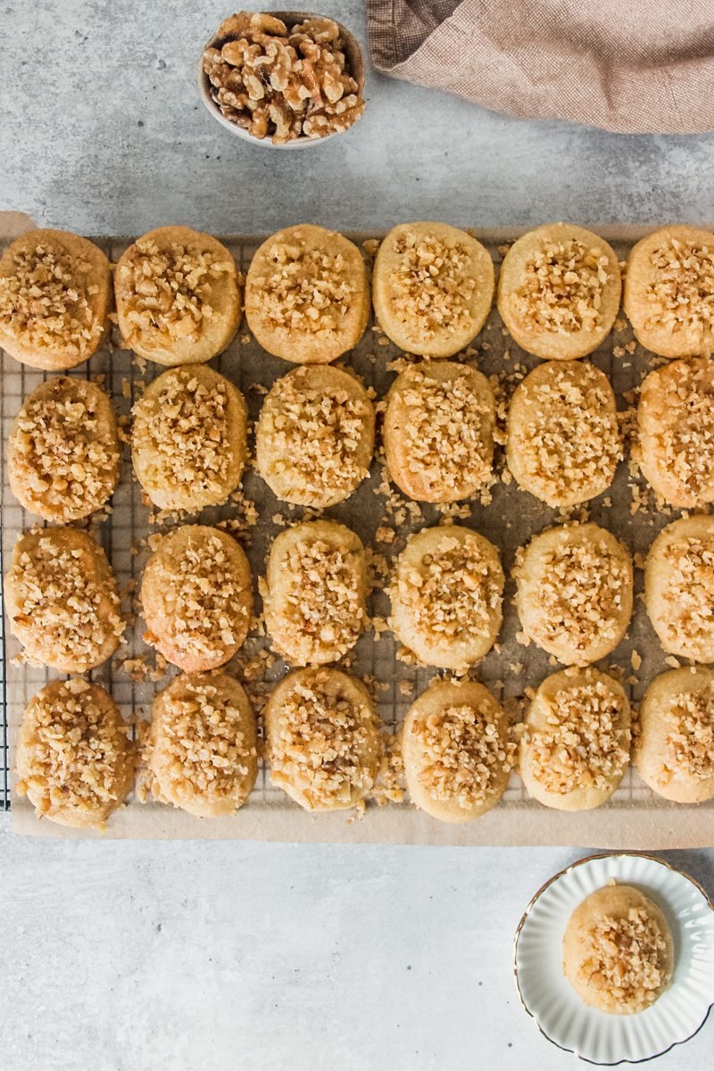 Melmakarona or Greek walnut christmas cookies sit on parchment paper on a cooling rack above a gray surface.