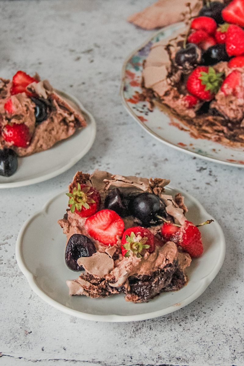 Individual servings of chocolate pavlova sits on white ceramic plates with the gooey center, chocolate cream and fruit topping visible on a light gray surface.