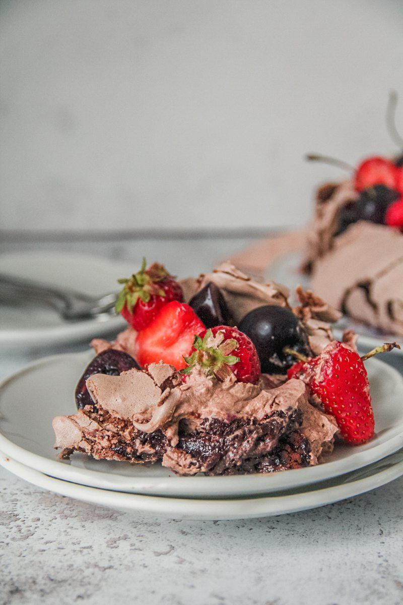 An individual serving of chocolate pavlova sits on a two white ceramic plates with the gooey center, chocolate cream and fruit topping visible on a light gray surface.