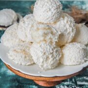 Austrian coconut meringue cookies sit on a white fluted ceramic plate on a wooden cake stand with one cookie sitting halved with the fluffy soft interior visible. Above a green and white surface.