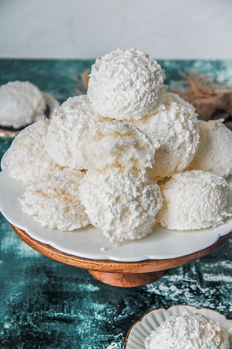 Austrian coconut meringue cookies sit on a white fluted ceramic plate on a wooden cake stand with one cookie sitting halved with the fluffy soft interior visible. Above a green and white surface.