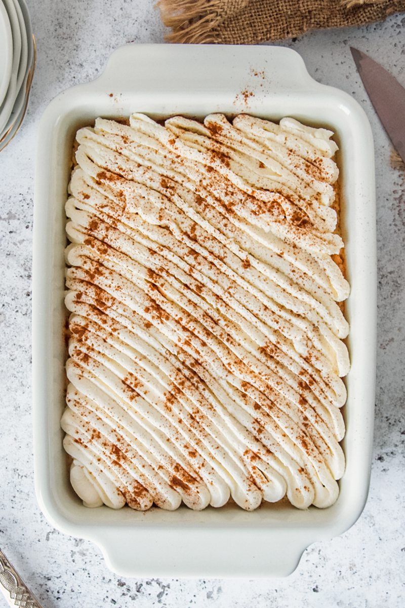 Tres Leches cake sits topped with whipped cream and a sprinkle of ground cinnamon on top in a rectangle white baking dish on a light gray surface.